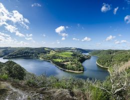Panoramablick auf einen Fluss, der sich durch eine grüne Hügellandschaft schlängelt, unter einem blauen Himmel mit weißen Wolken.