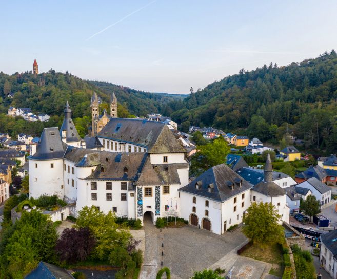 Luftaufnahme von Schloss Clervaux in Luxemburg, umgeben von Wald und Stadtgebäuden.